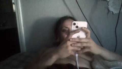 Media: A dimly lit video of a woman lying on her back, taking a selfie with her iPhone. She has light skin, long brown hair, and is wearing a black strapless top. The background is a plain, light-colored wall.