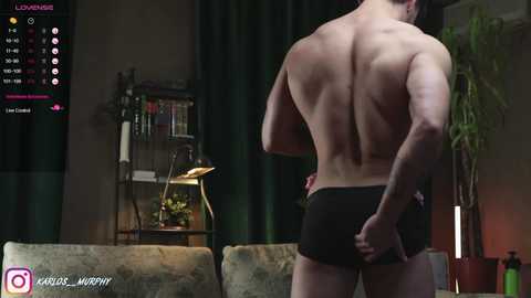 Media: Video of a muscular man with a tan back wearing black boxer briefs, standing in a dimly lit living room with green curtains, a bookshelf, and a plant.