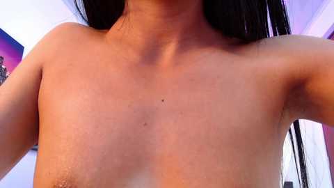 Media: Video of a topless, fair-skinned person with long black hair, revealing a mole on the chest. Background includes purple and white walls with indistinct objects.