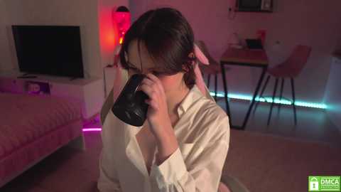 Media: A video of an Asian woman with medium brown hair in a white robe, drinking from a black mug in a dimly lit, modern bedroom with pink and purple LED lighting.