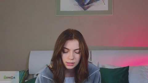 Media: Video of a young woman with long brown hair, wearing a denim jacket, sitting on a couch with green and white pillows, against a beige wall with a framed abstract art piece above.