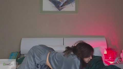 Media: Video of a woman in a blue nightgown lying on a bed with white headboard, surrounded by green sheets and red lighting, in a beige room with abstract art.