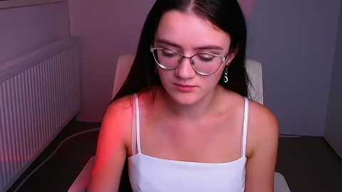 Media: Video of a young woman with straight black hair, wearing glasses and a white spaghetti-strap top, seated indoors with a red light casting a soft glow.