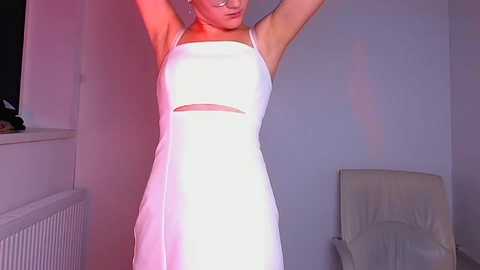 Media: Video of a fair-skinned woman with a slim build, wearing a white, sleeveless, halter-neck dress with a cutout midriff, standing in a minimalist room with a radiator, chair, and window on the left.