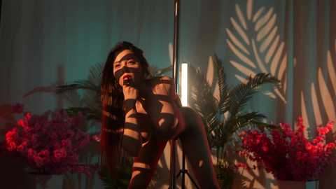 Media: Video of a naked woman with long dark hair, leaning over a pole, surrounded by vibrant red flowers and green ferns.