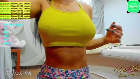 Media: Video of a fit woman with long black hair, wearing a tight yellow crop top and colorful shorts, standing in a bright, modern room.