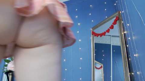 Media: Video of a woman lifting her pink plaid skirt, revealing her bare buttocks, in a room with blue walls, heart garland, and mirror.