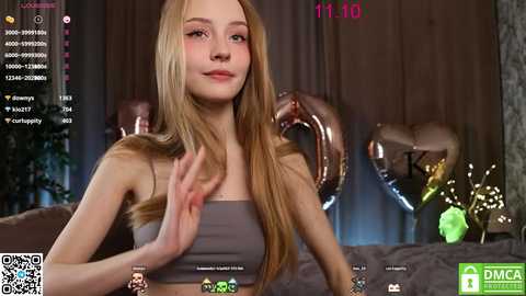 Media: Video of a young Caucasian woman with long, straight, light brown hair, wearing a gray sports bra, smiling while seated on a couch. The background features a dimly lit room with metallic heart balloons.