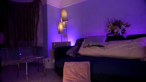 Media: Video of a dimly lit, cozy bedroom with a dark blue sofa, white and beige pillows, a silver stool, and a tall, modern floor lamp with a purple glow.