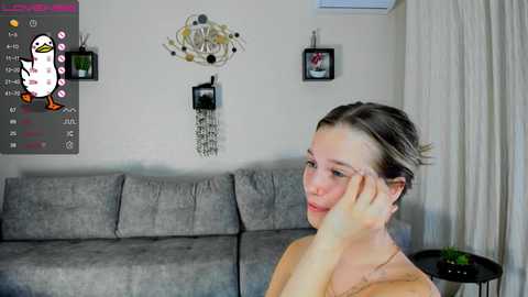 Media: Video of a young woman with fair skin and light brown hair, wearing glasses, touching her face, sitting on a gray sofa in a modern living room with white walls, gray curtains, and two framed pictures.