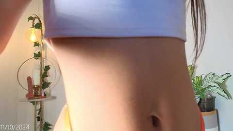 Media: Video of a slender, light-skinned woman with a flat stomach, wearing a white sports bra, taken indoors, showing her midsection. Background features a potted plant, a lamp, and a shelf.