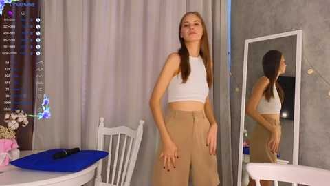 Media: Video of a young woman with long brown hair, wearing a white crop top and high-waisted beige pants, standing in a modern room with gray curtains, a mirror, and a white chair.