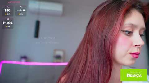 Media: Video of a young woman with long, reddish-brown hair, fair skin, and pink lipstick, seen from the side in a modern, blurred interior setting.