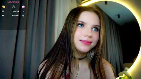 Media: Video of a young Caucasian woman with long, straight brown hair, blue eyes, and fair skin, wearing a black choker. She stands indoors with grey curtains and a circular mirror with warm lighting in the background.