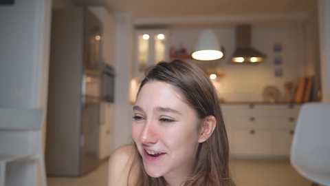 Media: Video of a smiling young woman with long brown hair, fair skin, and minimal makeup, in a modern kitchen with stainless steel appliances, white cabinets, and a hanging light fixture.