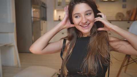 Media: Video of a smiling young woman with long brown hair, wearing a black sleeveless top, adjusting her hair in a modern kitchen with light-colored cabinets and a large fridge.