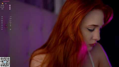 Media: Video of a fair-skinned woman with long, fiery red hair, wearing a white top, looking down. Background is blurred with purple and pink lighting, creating a moody atmosphere.