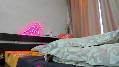 Media: Video of a modern bedroom with a neon pink heart sign on a white wall, a black headboard, and a messy bed with gray patterned sheets and a colorful blanket.