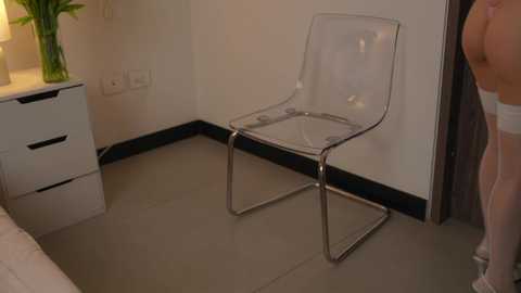 Media: Video of a modern, minimalist bedroom with a clear acrylic chair, a white dresser, and a bedside lamp on a beige carpet.