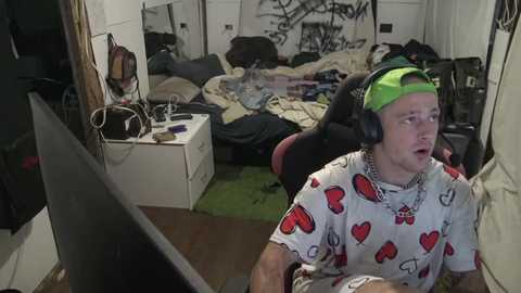 Media: A video of a young man with a green headband, wearing a white T-shirt with red heart patterns, sitting in front of a computer in a cluttered bedroom.