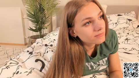 Media: Video of a young, fair-skinned woman with long, straight brown hair, wearing a green graphic tee, lying on a bed with a white comforter featuring black cat illustrations. Background includes a potted plant and a white radiator.