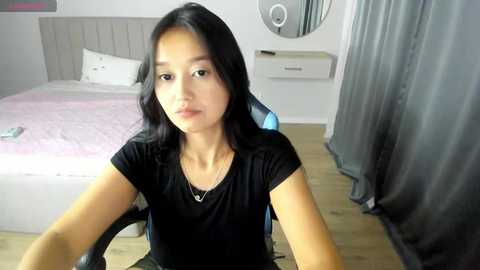 Media: Video of a young Asian woman with long black hair, wearing a black t-shirt, sitting in a blue gaming chair in a minimalist bedroom with a pink bed and gray curtains.