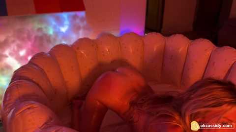 Media: A video featuring a nude woman with blonde hair and fair skin lying on a pink inflatable bed, illuminated by vibrant neon lights.