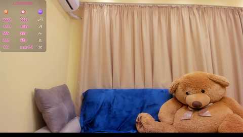 Media: Video of a cozy room with beige walls, a plush teddy bear on a blue couch, and sheer curtains, featuring a digital clock display and an air conditioner unit.