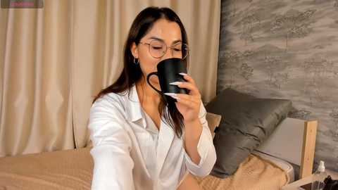 Media: Video of a young woman with long brown hair, glasses, white button-up shirt, drinking coffee in a modern bedroom with beige curtains, gray wallpaper, and a gray bed.