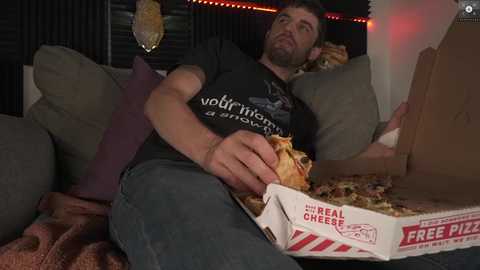 Media: Video of a man in a dark T-shirt, jeans, and glasses, reclining on a couch, eating pizza from a \"Free Pizzas\" box, with a blanket and a pillow nearby, in a dimly lit living room.