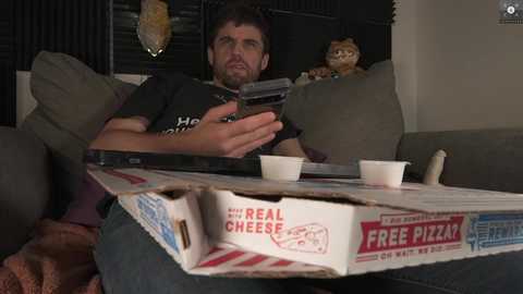 Media: A video shows a bearded man in a black T-shirt, holding a pizza box with a \"Real Cheese Pizza\" label, sitting on a couch with a stuffed animal and a remote control.