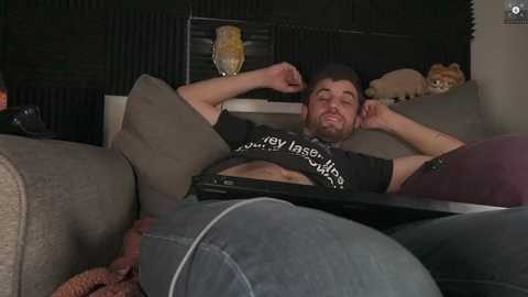 Media: Video of a bearded, muscular man in a black T-shirt and jeans, lying on a sofa, with a relaxed expression. Background includes a radiator, cushions, and a stuffed animal.