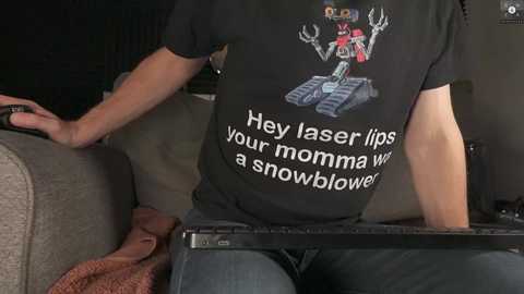 Media: A video shows a person wearing a black t-shirt with a LEGO minifigure and snowblower graphic. They are sitting on a couch, holding a controller, with a laptop on their lap. The background is dimly lit.