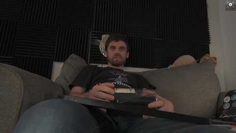 Media: Video of a man with a beard and headphones, wearing a dark T-shirt, reclining on a gray couch with black acoustic foam panels in the background.