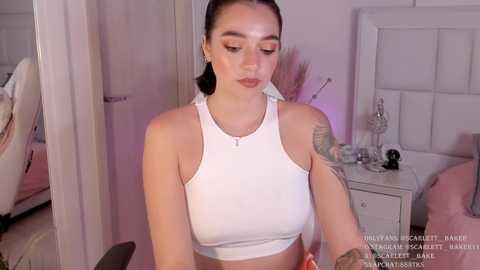 Media: A video of a young woman with light skin and dark hair tied back, wearing a white crop top, sitting in a modern, minimalist bedroom with white furniture and a bed.