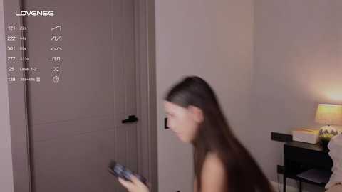 Media: Video of a young woman with long dark hair, possibly Asian, in a dimly lit bedroom. She holds a smartphone, standing near a closed door and a nightstand with a lamp.