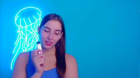 Media: Video of a young woman with straight brown hair, wearing a blue tank top, holding a lighter against a glowing neon jellyfish on a bright blue background.