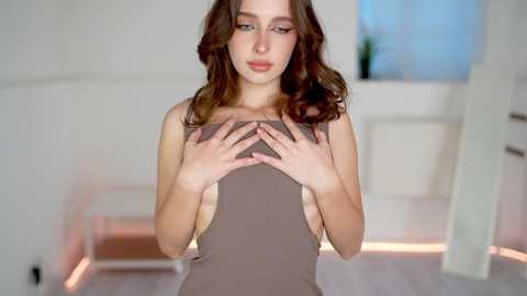 Media: Video of a young Caucasian woman with long, wavy brown hair, wearing a sleeveless, light brown dress, covering her chest with her hands, standing indoors in a minimalist setting with white walls and furniture.