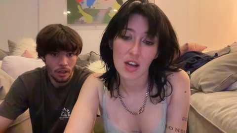 Media: A video shows a young, pale-skinned woman with dark hair and tattoos, wearing a tank top, sitting on a couch. A man with messy hair, in a T-shirt, looks at her. Background includes a colorful abstract painting and pillows.