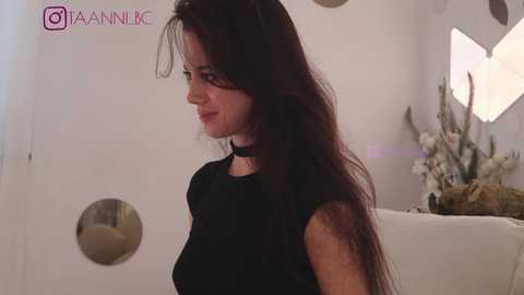 Media: Video of a woman with long dark hair, wearing a black top, standing indoors against a white wall with circular designs and a floral arrangement.