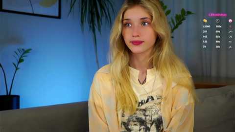 Media: Video of a young blonde woman with long hair, wearing a graphic T-shirt, sitting on a couch in a dimly lit living room with plants and framed artwork in the background.