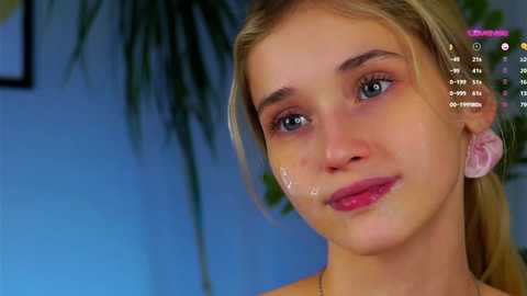 Media: Video of a young blonde woman with light skin, blue eyes, and pink lips, wearing a simple necklace. Background shows a blurred potted plant and blue wall.