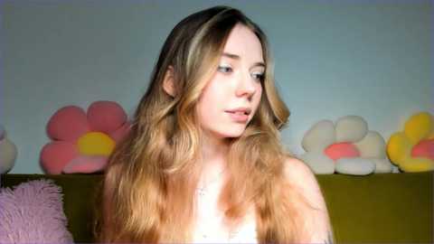 Media: A video of a young woman with long, wavy, light brown hair, wearing a white top, standing in a room with a light blue wall adorned with large, pink and yellow flower-shaped balloons.