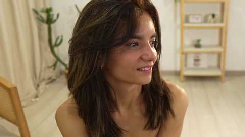 Media: Video of a young, light-skinned woman with long, dark brown hair, wearing a subtle smile, seated in a bright, minimalistic room with beige curtains, wooden furniture, and potted plants.