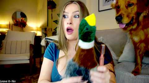 Media: Video of a blonde woman with a surprised expression wearing a blue top, holding a yellow duck costume, in a cozy living room with a brown sofa and a dog.