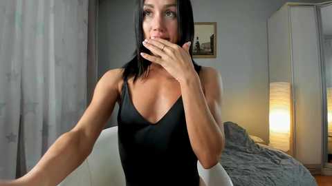 Media: Video of a fit, light-skinned woman with long black hair, wearing a black V-neck top, covering her mouth in surprise, seated on a white couch in a softly lit, minimalist bedroom.