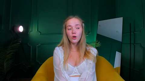 Media: Video of a blonde woman with long hair, wearing a sheer white lace top, sitting in a mustard-yellow chair, in a dimly lit, green-paneled room with a spotlight and ferns.