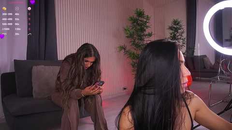 Media: Video of a young woman with long black hair, in a light brown coat, leaning on a dark gray sofa, looking at her phone.