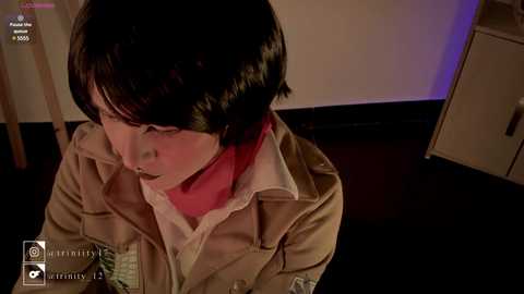 Media: Video of a young Asian woman with short black hair, wearing a beige jacket over a white shirt and red scarf, sitting on a dark floor, with a dimly lit room in the background.