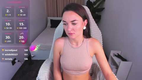 Media: A video shows a slender, fair-skinned woman with dark hair, wearing a beige crop top and white earrings, in a modern bedroom with a white bed and green plant.